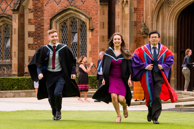Three graduates (2 males 1 female) run towards camera in their robes with Lanyon building in backgrouns